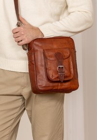 Sac à bandoulière en cuir marron avec un rabat frontal, fermeture par boucle et une bandoulière. Présente des coutures visibles pour un détail supplémentaire.