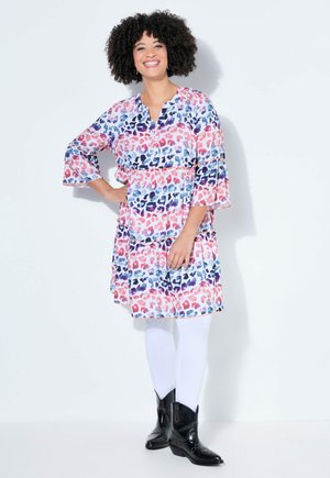 Lächelnde Frau mit lockigem Haar, die ein buntes Leopardenmusterkleid, weiße Leggings und schwarze Cowboy-Stiefel trägt, stehend mit einer Hand in der Hüfte.