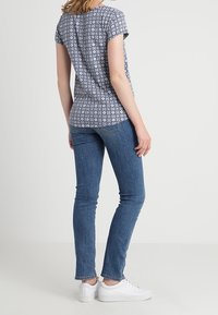 Camisa de manga corta de patrón floral azul combinada con jeans ajustados azules y zapatillas blancas, vista desde atrás.