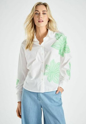 Witte overhemd met grote groene bloemenborduursels op de mouwen; gecombineerd met lichtblauwe wijde pijpen. Soepele katoenen textuur.