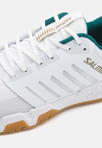 Hvid sportsko med mesh-overdel, teal detaljer og gyldent branding. Bemærkelsesværdig gummisål med hexagonal mønster og plastoverlag for støtte.