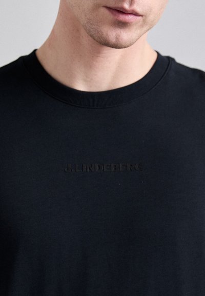 Camiseta de algodón negra con cuello redondo que presenta un logo de la marca "J.LINDEBERG" sutilmente grabado en el pecho. Textura suave.
