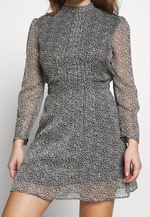 Frau trägt ein schwarz-weißes, langärmliges Minikleid mit Blumenmuster, transparenten Ärmeln und hohem Kragen.