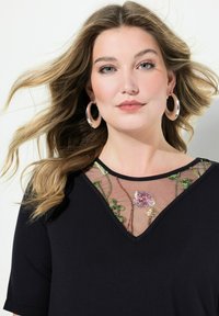 Top negro con escote en V y bordado floral transparente en el cuello. La modelo tiene el cabello ondulado y lleva pendientes de aro grandes.