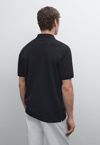 Černé polo tričko s krátkými rukávy, z bavlněného materiálu, jednobarevné, s postranními rozparky na lemu a volným střihem. Model je zobrazen zezadu.
