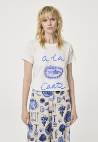 Roomwit kort-sleeve T-shirt met blauwe tekst "a la Carte" en vis afbeelding. Gecombineerd met beige broek met blauwe voedselgerelateerde illustraties.