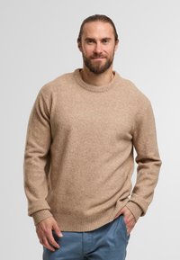 Maglione beige in maglia con colletto a girocollo, polsini e orlo a coste, realizzato in un materiale morbido, indossato sopra pantaloni azzurri chiari.