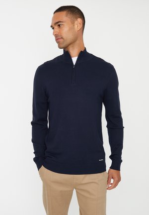 Pull bleu marine en maille avec zip quart, en tissu tricoté, et finitions côtelées aux manches et à l'ourlet. Présente une petite étiquette de marque en bas.