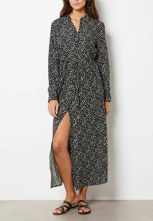 Robe maxi à pois noirs et blancs avec manches longues, col à revers, taille à cordon et fente latérale haute jusqu'à la cuisse.