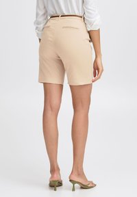 Beige, letisított rövidnadrág, lapos elejével, két hátsó zsebével és sima, egységes textúrával, zöld magassarkú szandálokkal párosítva.