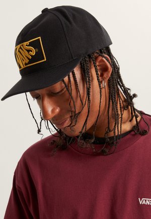 Svart keps med en guld "VANS"-logotyp på framsidan, med en böjd skärm. Bäraren har flätat hår och är klädd i en vinröd sweatshirt.