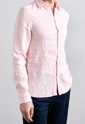 Hombre con camisa rosa clara de manga larga abotonada con cuello y pantalones azul marino, de pie con una mano en el bolsillo.