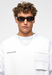 Lunettes de soleil de forme rectangulaire avec des verres foncés. Sweat-shirt gris avec une poche cousue et "Wonders" brodé en noir. Deux chaînes en argent.