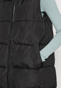 Zwarte gewatteerde bodywarmer met een ritssluiting aan de voorkant, met een geborduurd ontwerp, twee zijzakken en een comfortabele pasvorm, gedragen over een lichtblauw ribbelshirt.