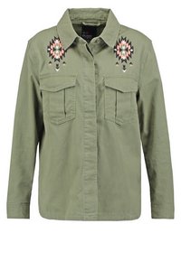 Camisa chaqueta en verde oliva de mezcla de algodón con dos bolsillos en el pecho. Presenta un colorido bordado geométrico en los hombros. Cuello plano, mangas largas.