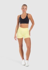 Zwarte sportbeha met een ronde hals en geplooide details, gecombineerd met hoge, gele rekbare short. Model draagt witte sneakers.