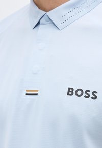 Ανοιχτό μπλε πόλο πουκάμισο με διάτρητο γιακά, τρία κουμπιά και το λογότυπο BOSS δίπλα σε ένα μικρό μαύρο, λευκό και καφέ ριγέ έμβλημα.