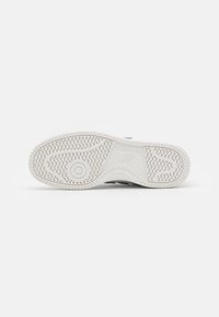 Suela blanca con agarres texturizados; la entresuela presenta una superficie lisa. Acentos en negro y gris cerca del talón. Marca clara visible.