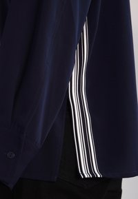 Camisa de color navy con mangas largas, que presenta una abertura lateral y rayas verticales blancas y negras en contraste a lo largo del dobladillo para un detalle adicional.