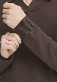 Tamno smeđi pleteni pulover s V-izrezom i rebrastim manžetama. Tkanina ima teksturirani uzorak, prikazujući usko prileganje na zapešćima.