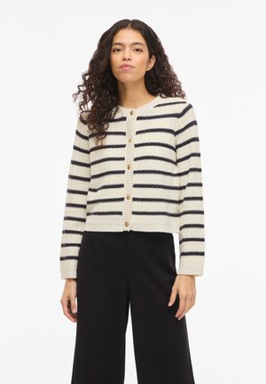 Moteris su garbanotais plaukais, vilkinti kremine megztinio kardigano su juodomis horizontaliomis juostelėmis ir medinėmis sagutėmis, derinama su juodomis pločio kojų kelnėmis.