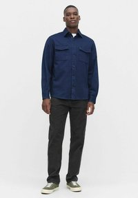 Marineblauw overhemd met knopen, voorzien van twee borstzakken, gecombineerd met zwarte broek en groene sneakers. Het shirt heeft een kraag en lange mouwen.