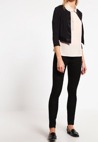 Schwarze taillierte Jacke mit Dreiviertelärmeln über einer hellbeigen gefalteten Bluse, kombiniert mit schwarzen Leggings und schwarzen Slippern.