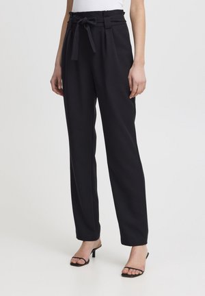 Kvinde iført sort buks med højtaljede og et bindebælte af stof samt sorte sandaler med hæl og stropper, stående mod en ensfarvet baggrund.