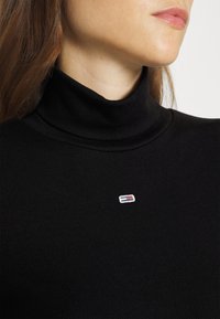 Schwarzer gerippter Rollkragenpullover mit einem kleinen, mehrfarbigen gestickten Logo auf der Brust. Weicher Stoff, figurbetonendes Design.