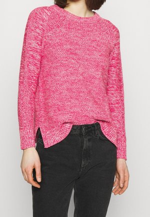 Personne portant un pull rose en tricot à manches longues et un jean noir taille haute, debout devant un fond clair uni.