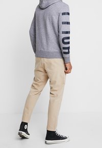 Grå hoodie med marinblå tryckt text på ärmen, i kombination med beige byxor och svarta högsneakers. Enkel design och avslappnad passform.