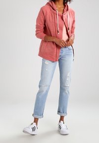 Sudadera roja con cordones, camiseta rosa debajo, vaqueros desgastados azul claro y zapatillas blancas con rayas negras. Conjunto casual mostrado.