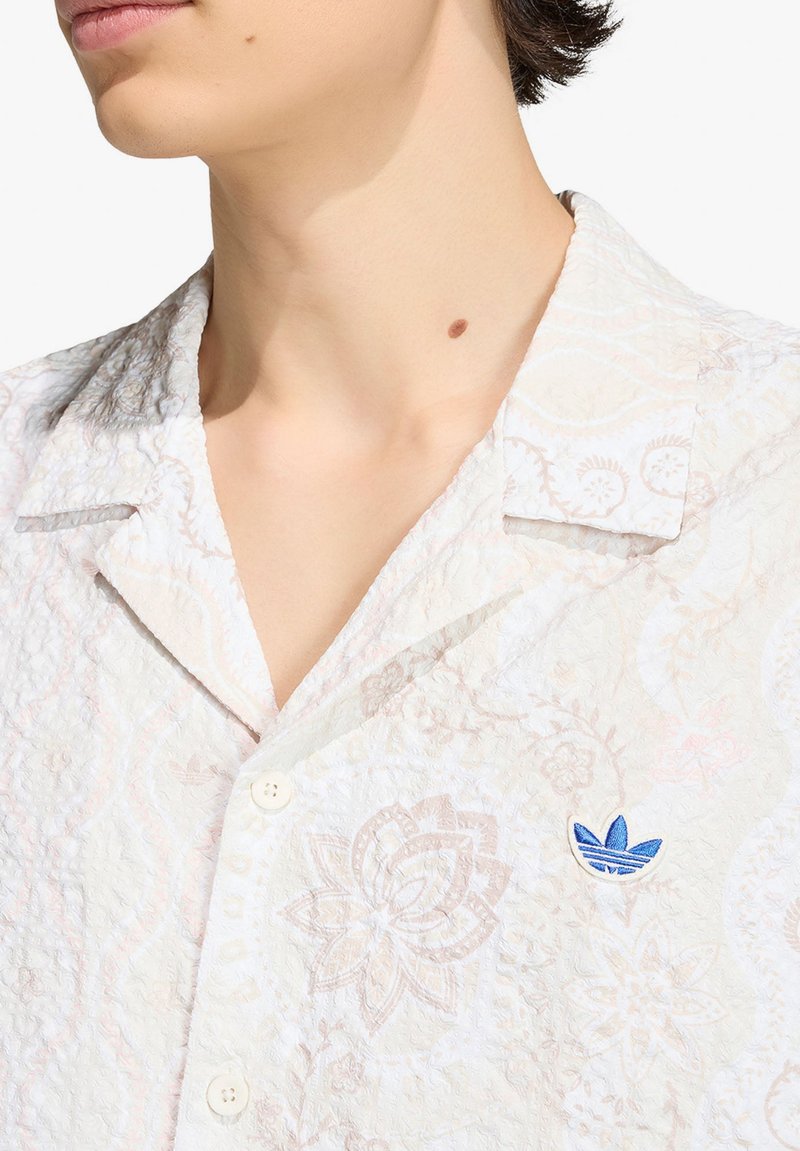 Persona che indossa una camicia bianca con bottoni e trama, con motivi floreali pallidi e un logo Adidas blu sul petto, mostrando collo e mento.