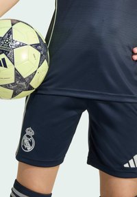 Blå sportshorts med vita Adidas-ränder och Real Madrid-logotyp. Håller en gul och svart UEFA Champions League-fotboll.