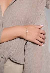 Złota bransoletka z sercem i przezroczystymi cyrkoniami, noszona na nadgarstku o neutralnym odcieniu skóry. Widoczny teksturowany jasnobrązowy top.