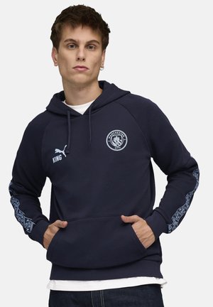 Mørkeblå hoodie med kængurulomme, justerbar snoretrækshætte og mønstrede detaljer på ærmerne. Har Manchester City logo og teksten "KING".