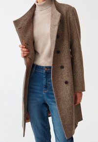 Abrigo marrón de pata de gallo con grandes botones negros, bolsillos laterales y un interior suave. Lleva puesto sobre un jersey de cuello alto beige y vaqueros azules.