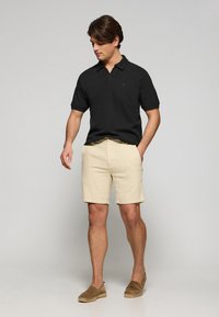 Svart pikétröja med korta ärmar, beiga shorts och bruna espadrillos. Enkelt mönster med krage och subtil logotyp på tröjan.