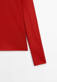 Manche longue rouge d'un vêtement sur fond blanc, montrant une texture de tissu lisse et une couture propre à l'ourlet.