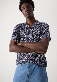 Camisa de mangas curtas em azul-marinho com padrão abstrato em branco. Apresenta um colarinho, botões e um corte estilo casual, combinada com jeans de denim azul claro.