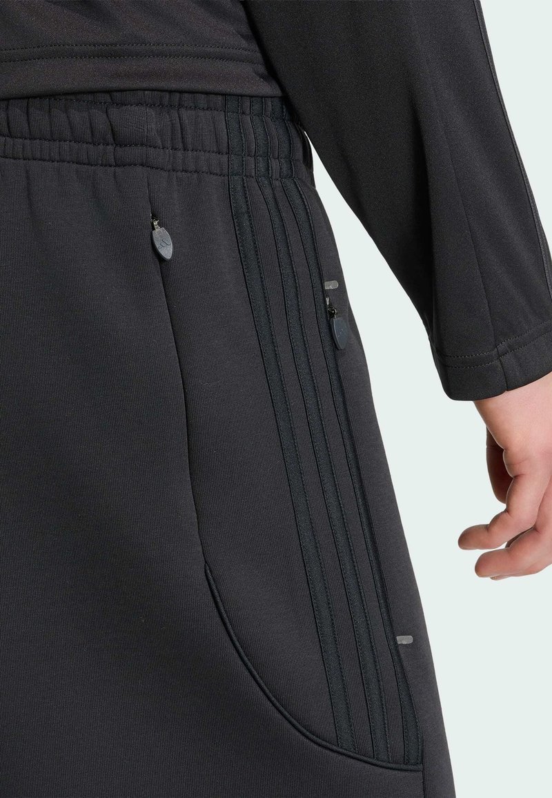Primo piano di una persona che indossa pantaloni sportivi neri con tasche con cerniera e dettagli a strisce laterali, mano rilassata al fianco.