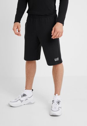 Man draagt zwarte EA7 shorts, zwarte top met lange mouwen, witte sokken en wit-grijze sportschoenen, staande tegen een witte achtergrond.
