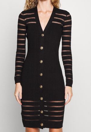 Robe noire côtelée avec manches longues transparentes à rayures et profond décolleté en V. Des boutons dorés ornent le devant, rehaussant le design.