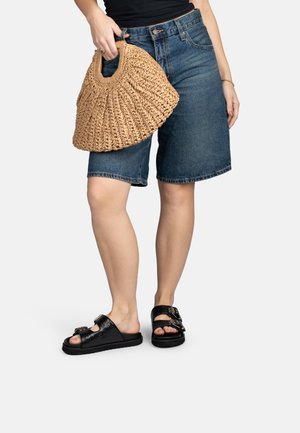 Donna che indossa pantaloncini bermuda di jeans, sandali neri a ciabatta con fibbie, tiene in mano una borsa intrecciata color cuoio, in piedi su uno sfondo bianco.