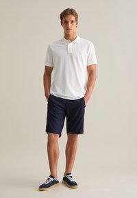 Camisa polo blanca de manga corta combinada con pantalones cortos azul marino y zapatillas azul marino. La tela es suave; los pantalones cortos tienen dobladillos enrollados para un look casual.