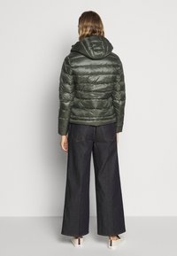 Žena v tmavě zelené prošívané bundě s kapucí, širokých tmavých džínách a bílých teniskách se pruhovaným detailem, stojící zády.