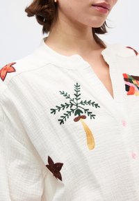 Femme portant un chemisier blanc texturé avec des motifs floraux et feuillages brodés colorés près de l'épaule et de la poitrine.