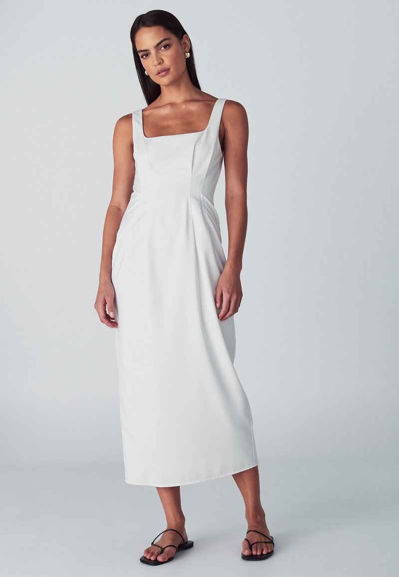 Žena v bezrukávových bielych midi šatách so štvorcovým výstrihom a čiernymi plochými sandálmi, stojiaca pred jednoducho svetlošedým pozadím.