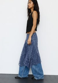 Hosszú hajú nő, aki ujjatlan fekete mellényt, réteges kék csipkés szoknyát széles farmernadrág felett és fekete hegyes cipőt visel, oldalra sétál.