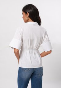 Blusa bianca a maniche corte con vita cincta, caratterizzata da un colletto e tessuto testurizzato. Abbinata a jeans blu a vita alta.